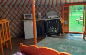 Interior de una yurta con muebles coloridos, pequeña cocina y puerta abierta con vista al césped verde.