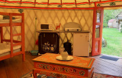 Interior de una yurta tradicional con litera, cocina pequeña, cofre decorado y puertas al jardín.