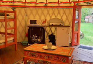 Interior of a traditional yurt with bunk bed, kitchenette, ornate chest, and doors opening to grass.