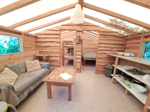 Indvendig udsigt over en hyggelig træhytte med sofa, bord, køkkenområde og sovepladser i baggrunden.