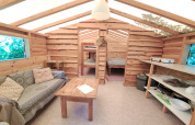 Innenansicht einer gemütlichen Holzhütte mit Sofa, Tisch, Küchenbereich und Schlafplätzen im Hintergrund.