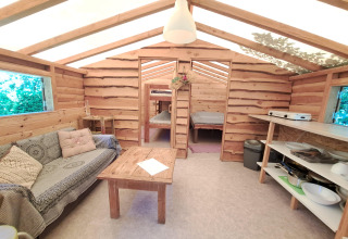 Vue intérieure d'une cabane en bois chaleureuse avec canapé, table, coin cuisine et espaces de couchage au fond.