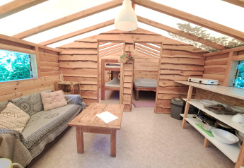 Vue intérieure d'une cabane en bois chaleureuse avec canapé, table, coin cuisine et espaces de couchage au fond.