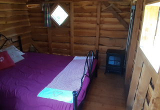 Chambre d’une cabane perchée avec murs en bois, lit violet et poêle au camping Ushuaïa Villages Le Viaduc.
