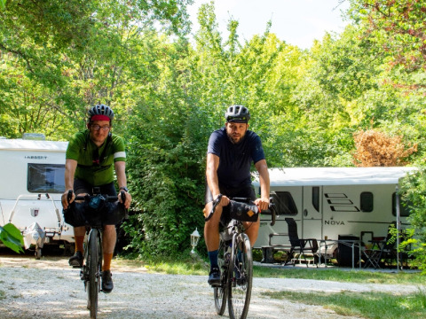 Deux cyclistes casqués roulent sur un camping verdoyant avec des caravanes et des arbres à l'arrière-plan.