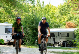 Deux cyclistes casqués roulent sur un camping verdoyant avec des caravanes et des arbres à l'arrière-plan.