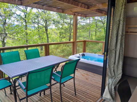 Terrazza in legno al Camping Le Luberon con tavolo, sedie verdi e idromassaggio, vista sul bosco.