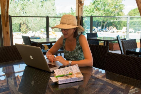 Femme avec chapeau de soleil travaillant sur portable à une table en verre au camping Le Luberon, Provence.