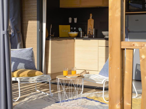 Terrazza soleggiata al Lodge del Campeggio La Nouvelle Croze, Francia, con sedie, cuscini e succo.