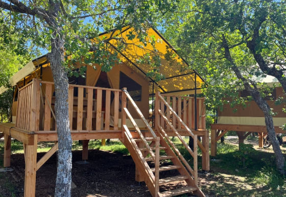 Safari-Zelt namens Bungalowtent auf einer erhöhten Holzterrasse, umgeben von grünen Bäumen.