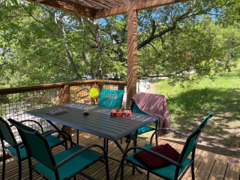 Terrazza all’aperto del Cottage «Pantaïa» presso il Campeggio Le Luberon, con tavolo e sedie tra gli alberi.