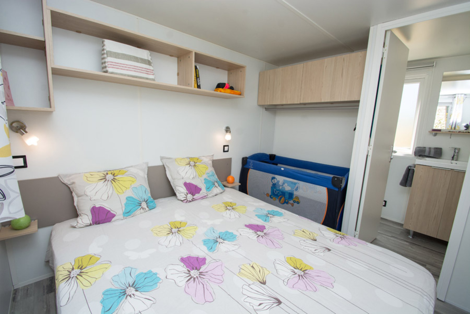 Slaapkamer in Cottage Romarin op Camping Le Luberon in Frankrijk met bloemendekbed en babybed.