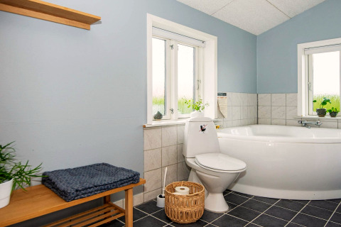 Lyst badeværelse med hjørnebadekar, toilet, træbænk og grønne planter i et feriehus i Haderslev, Danmark.