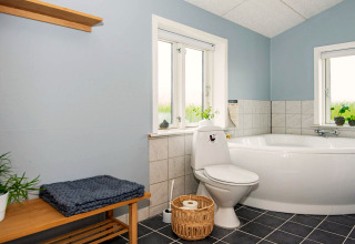 Salle de bain lumineuse avec baignoire d’angle, toilettes, banc en bois et plantes à Haderslev, Danemark.