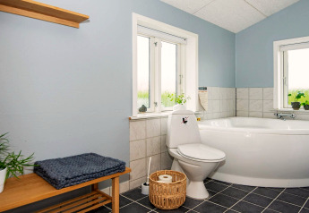 Salle de bain lumineuse avec baignoire d’angle, toilettes, banc en bois et plantes à Haderslev, Danemark.