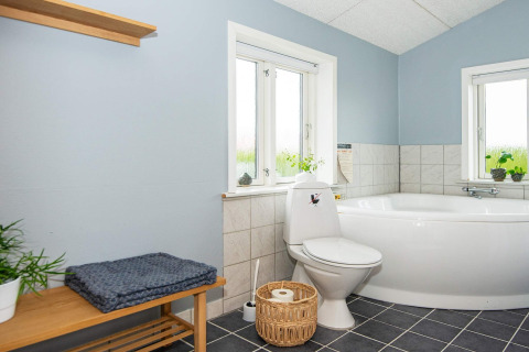Salle de bain lumineuse avec baignoire d’angle, toilettes, banc en bois et plantes à Haderslev, Danemark.