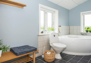 Salle de bain lumineuse avec baignoire d’angle, toilettes, banc en bois et plantes à Haderslev, Danemark.