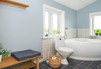 Salle de bain lumineuse avec baignoire d’angle, toilettes, banc en bois et plantes à Haderslev, Danemark.