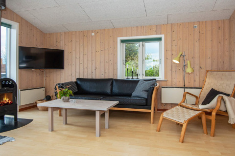 Salon d'une maison de vacances à Haderslev, Danemark, avec canapé noir, poêle et murs en bois.