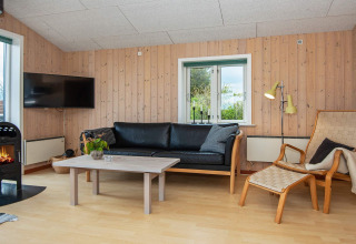 Salon d'une maison de vacances à Haderslev, Danemark, avec canapé noir, poêle et murs en bois.