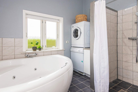 Salle de bain avec baignoire d’angle, douche, lave-linge, sèche-linge et fenêtre dans un gîte à Haderslev.
