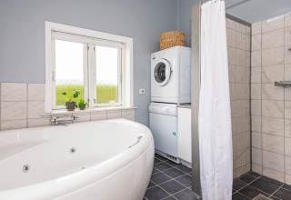 Salle de bain avec baignoire d’angle, douche, lave-linge, sèche-linge et fenêtre dans un gîte à Haderslev.