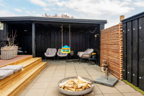 Coin salon extérieur avec murs noirs, fauteuils, brasero et balançoire dans une maison de vacances à Haderslev.