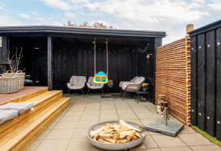 Coin salon extérieur avec murs noirs, fauteuils, brasero et balançoire dans une maison de vacances à Haderslev.