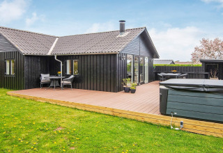 Maison de vacances noire avec grande terrasse et jacuzzi, située dans un cadre verdoyant à Haderslev, Danemark.