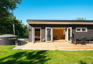 Sort feriehus med udendørs terrasse, spisebord, grøn have og spabad, beliggende i Glesborg, Danmark.