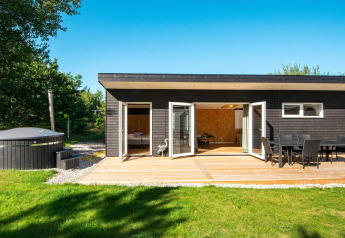 Casa de vacaciones negra con terraza, comedor exterior, césped y jacuzzi en Glesborg, Dinamarca.