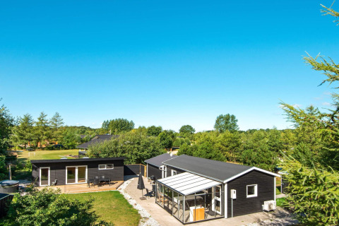 Sommerhus i Glesborg, Danmark, omgivet af træer, med terrasse, udestue og klar blå himmel ovenover.
