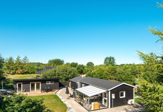 Sommerhus i Glesborg, Danmark, omgivet af træer, med terrasse, udestue og klar blå himmel ovenover.