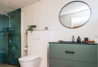 Moderne badeværelse i feriehus i Glesborg, Danmark, med grøn brusekabine, toilet og rund spejl.
