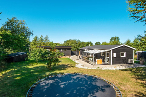 Sort feriehus i Glesborg, Danmark, med stor have, solterrasse og trampolin under blå himmel.