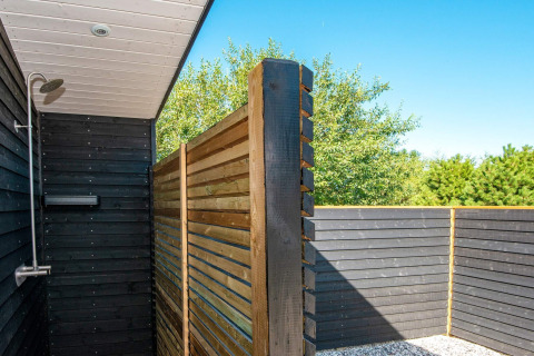 Udendørs bruser ved ferieboligen med trævægge og udsigt til grønne træer under en blå himmel.