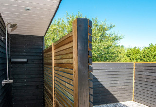 Udendørs bruser ved ferieboligen med trævægge og udsigt til grønne træer under en blå himmel.