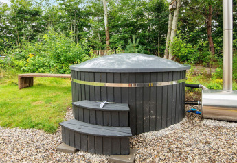 Bain à remous extérieur en bois noir avec bande métallique, posé sur gravier près d’un jardin arboré verdoyant.