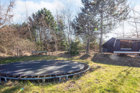 Trampolin og bænk i have ved feriebolig i Glesborg, Danmark med træer og hytte i baggrunden.