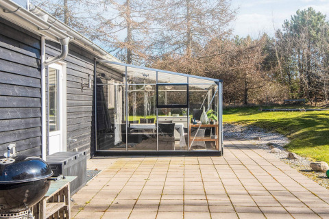 Solrig terrasse og glashave ved feriebolig i Glesborg, Danmark, omgivet af natur og træer.