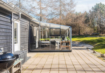 Solrig terrasse og glashave ved feriebolig i Glesborg, Danmark, omgivet af natur og træer.
