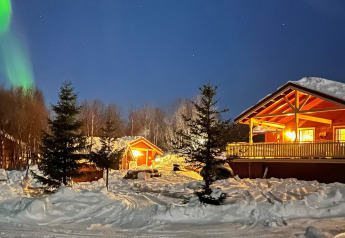 Besneeuwd chalet in Jarfjord, Zweden, ’s nachts verlicht met noorderlicht en sparrenbomen.