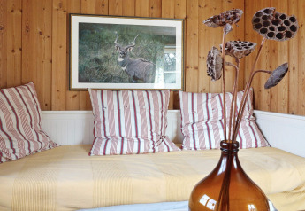 Acogedor rincón en chalet de madera con cojines a rayas, ropa de cama amarilla y jarrón marrón con flores secas.