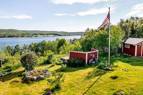 Vista de un chalet noruego en la ribera del río con bandera, cabañas rojas, vegetación y naturaleza verde.