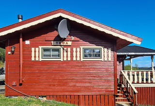 Rood houten chalet met satellietschotel, bord en veranda onder blauwe lucht in Jarfjord, Zweden.