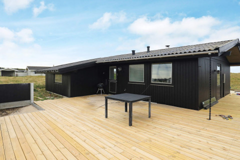 Sort sommerhus med træterrasse og have set udefra, Hjørring, Danmark, under en klar blå himmel.