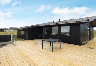 Zwart vakantiehuis met houten terras in Hjørring, Denemarken, bij zonnig weer en blauwe lucht.