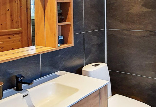 Baño moderno en una cabaña sueca con azulejos oscuros, lavabo, grifo negro, espejo y mueble de madera.