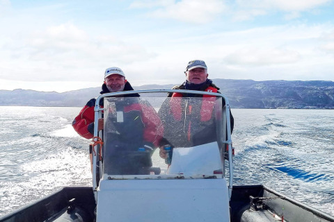 Twee mannen in rode jassen besturen een boot op het water met bergen op de achtergrond in Jarfjord, Zweden.