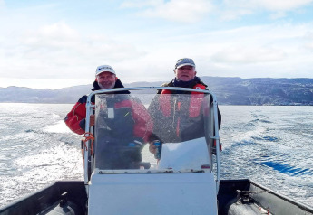 Dos hombres con chaquetas rojas conducen una lancha sobre el agua con montañas al fondo en Jarfjord, Suecia.